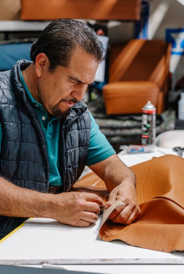 A man in a teal shirt and black vest works carefully with brown leather fabric on a table in a workshop, surrounded by tools and supplies.