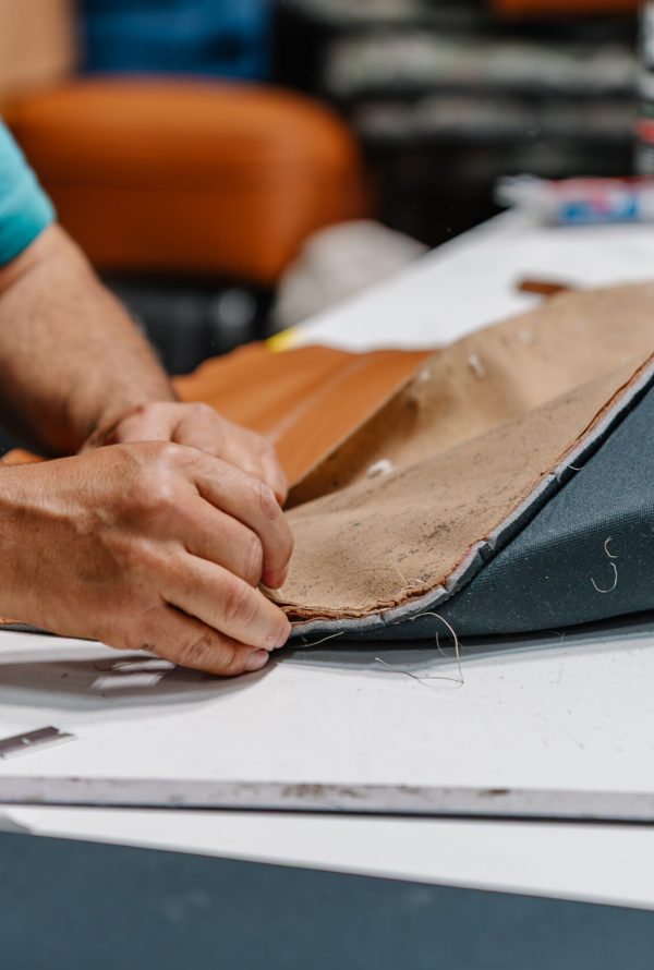 A person’s hands sewing fabric together on a work table, working on upholstery with tan and dark grey materials. Sewing tools and other materials are visible in the background.