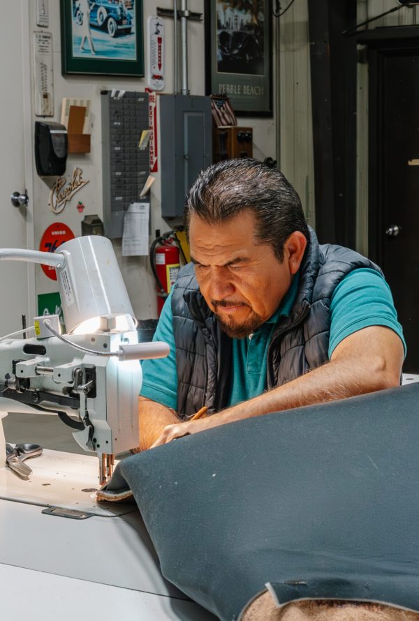 A man works intently at an industrial sewing machine in a workshop, stitching material on a worktable. Shelves, tools, and posters are visible in the background.