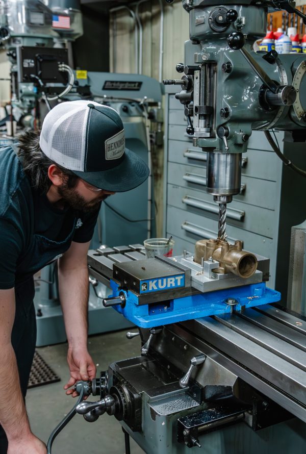 A man wearing a cap and apron operates a milling machine in a workshop, adjusting controls and working with a brass component clamped in a vise on the metal table.