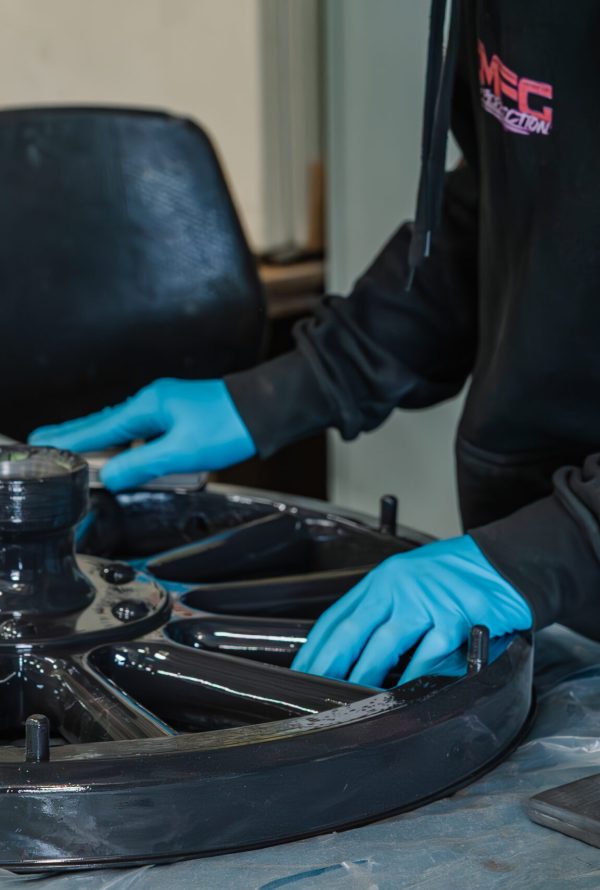 A person wearing blue gloves works on a large black metal wheel at a table, possibly cleaning or inspecting it. The person's face is not visible.