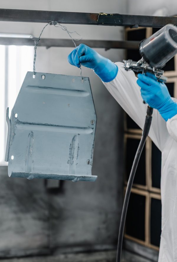 A person wearing protective gear and gloves uses a spray gun to paint a suspended metal part in an industrial setting, with a white mist in the air and bright windows in the background.