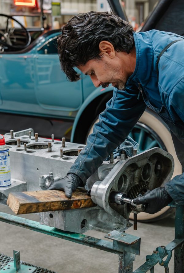 A man in a blue jumpsuit and black gloves works on a vintage car engine in a garage, with a classic blue car in the background and various tools and equipment around him.