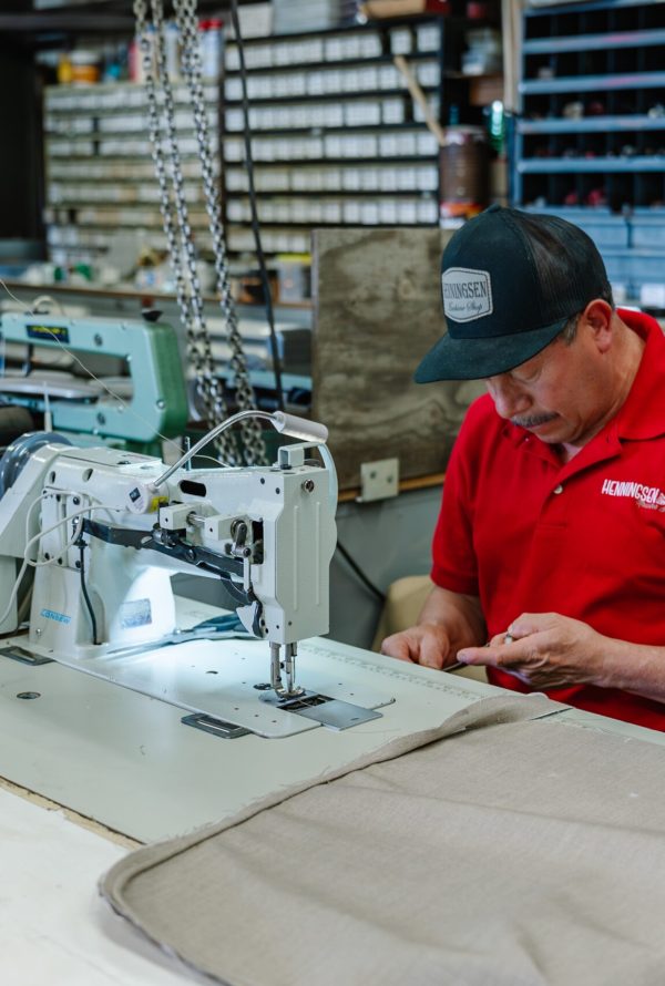 A man in a red shirt and black cap operates an industrial sewing machine in a workshop, stitching fabric with tools and equipment visible in the background.