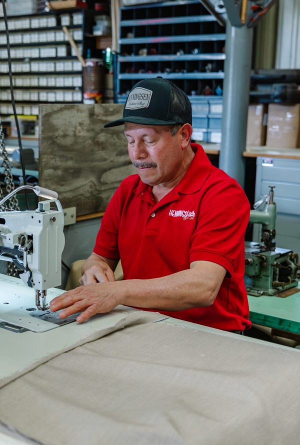 A man in a red shirt and black cap operates an industrial sewing machine, stitching light-colored fabric in a workshop with shelves and organized tools in the background.