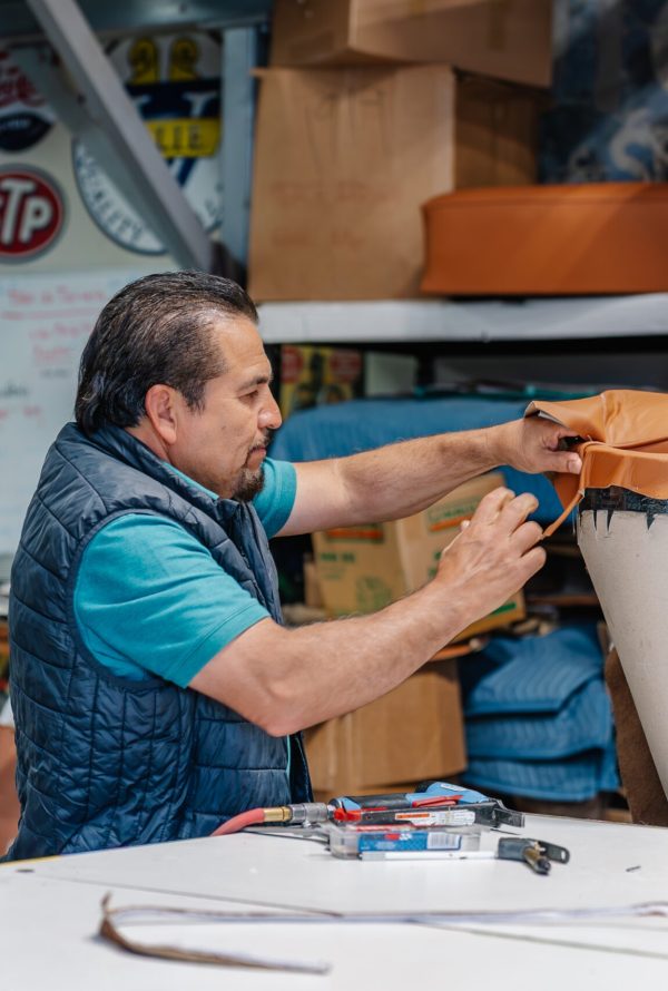 A man reupholsters an orange car seat in a workshop filled with tools, signs, and automotive memorabilia. He trims fabric while working at a cluttered table.