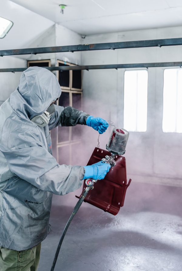 A person wearing protective clothing and gloves uses a spray gun to paint a metal object red inside a well-lit, ventilated workshop or spray booth.