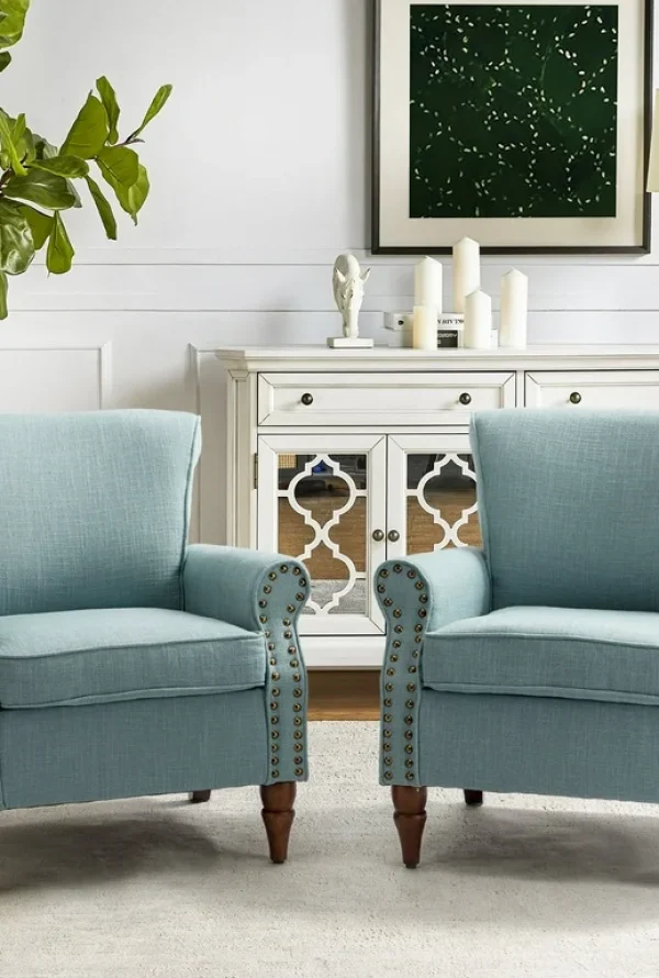 Two light blue armchairs with nailhead trim sit side by side in a living room with a white sideboard, decorative items, a blue lamp, a green potted plant, and a framed abstract artwork on the wall.