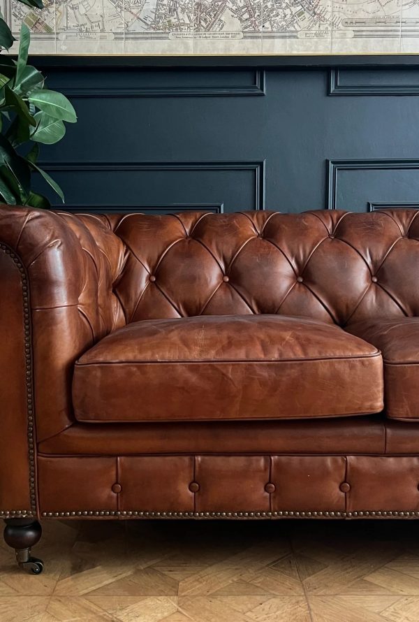 A brown leather tufted sofa with nailhead trim sits on a parquet floor against a dark paneled wall. A potted plant is on the left and a framed map hangs above.